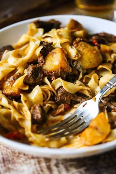 A close-up view of a white bowl that is filled with Hungarian goulash tossed with egg noodles and a portion of it missing.