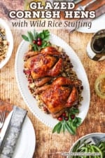 An overhead view of two glazed Cornish hens on a platter with wild rice stuffing flanked by a bowls of green beans and more stuffing.