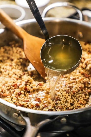 A person pouring chicken stock from a metal ladle into a large silver skillet filled with arborio rice and crispy pancetta.