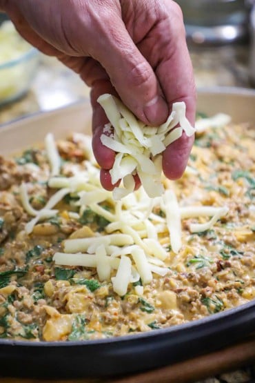 A person sprinkling shredded mozzarella over the top of a sausage, bean, and spinach dip in an oval baking dish.