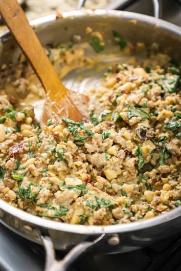 A front-on view of a large silver skillet fill with sautéd Italian sausage, white beans, pieces of sun-dried tomatoes, and wilted spinach.