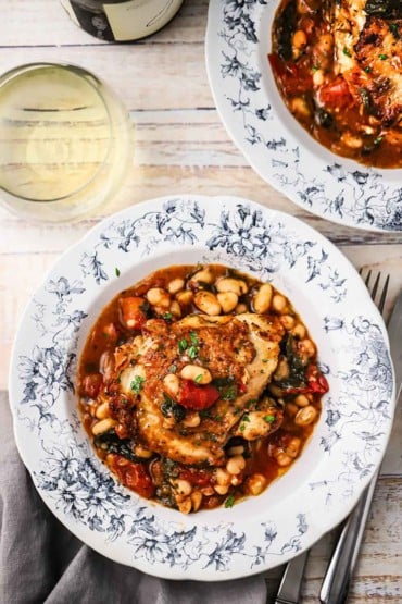 An overhead view of two antique dinner bowls filled with a serving of Tuscan chicken on top of a white bean, spinach, and tomato sauce.