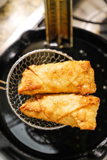 Two Philly cheesesteak egg rolls in a metal spider over a cast-iron skillet filled with hot oil.