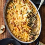 An overhead view of an oval baking dish filled with a partially eaten sausage, bean, and spinach dip with a silver spoon inserted into the side of the dip.