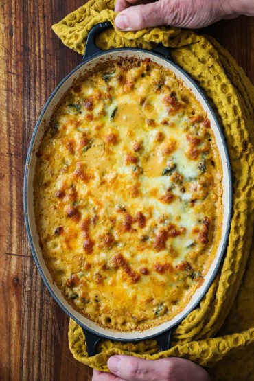 An overhead view of a person using a yellow kitchen towel to grip the handles of an oval baking dish filled with a sausage, beans, and spinach dip.