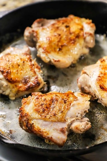 A straight-on view of four bone-in, skin-on chicken thighs that have been seared in butter in a cast-iron skillet.