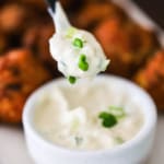 A small spoon being held up holding a spoonful of white remoulade topped with chopped scallions over a bowl of the same.