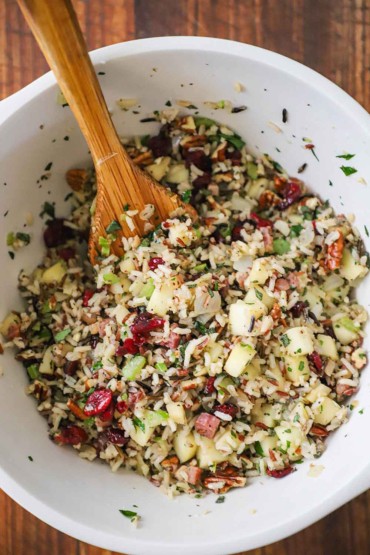 An overhead view of a white ceramic bowl filled with wild rice stuffing with pecans and cranberries as well as cubed apples, pancetta, and herbs.