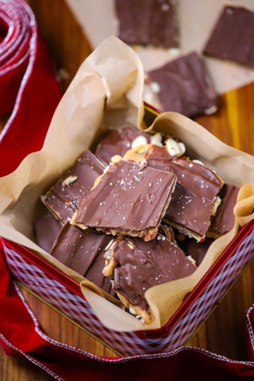 An overhead view of a Christmas candy tin filled with square pieces of chocolate toffee with red ribbon along the side of the tin.
