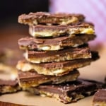 A close-up view of a stack of square chunks of chocolate toffee with almonds sitting on brown wax paper.