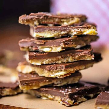A close-up view of a stack of square chunks of chocolate toffee with almonds sitting on brown wax paper.