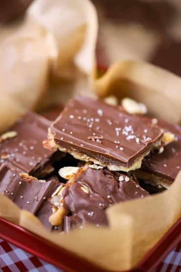 A close-up view of a pile of chocolate toffee with almond and sea salt in a festive red and white checkered Christmas tin.