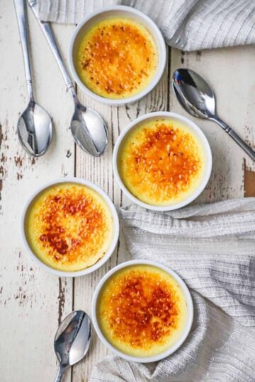 An overhead view of four white ramekins that are filled with lightly browned crème brûlée with several spoons scattered nearby.
