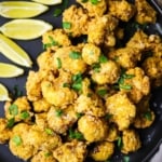 An overhead view of a black circular plate filled with a pile of fried cauliflower with lemon with a row of lemon wedges off to the side of the plate.