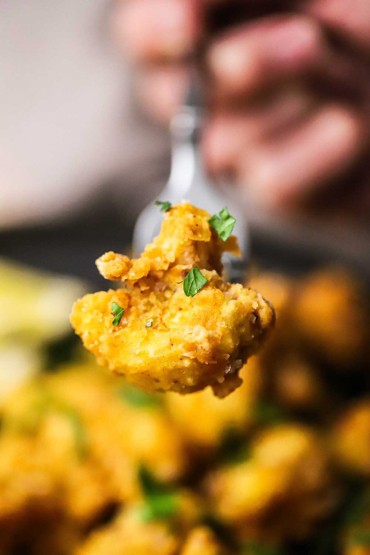 A close-up view of a piece of fried cauliflower with lemon that has been pierced with a fork and is being held up.