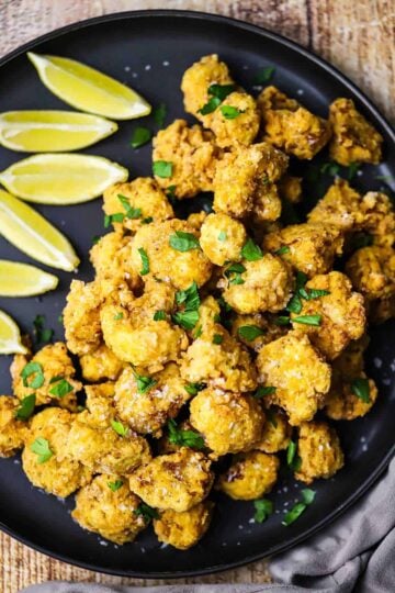 An overhead view of a black circular plate filled with a pile of fried cauliflower with lemon with a row of lemon wedges off to the side of the plate.