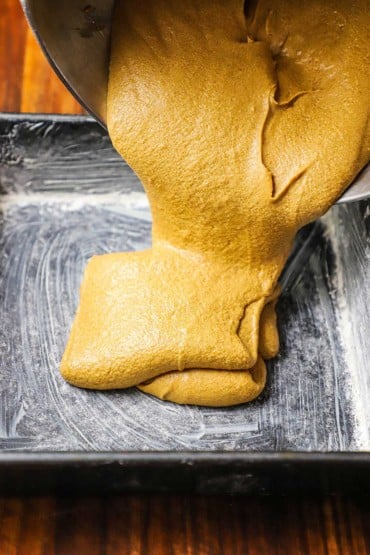 A golden gingerbread cake batter being poured into greased and floured square metal baking pan.