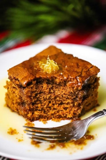 A straight-on view of a square piece of gingerbread cake with lemon sauce sitting on a white dessert plate with a bite out of it.