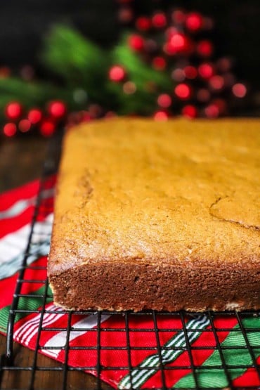 A whole square fully baked gingerbread cake sitting on a baking rack over a festive holiday place mat and in front of shiny red berries and green holly.