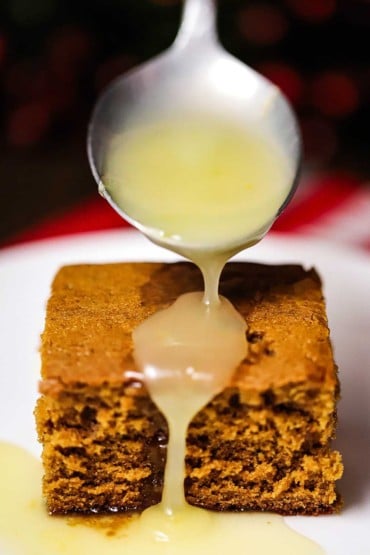 A large silver spoon being used to drizzle a lemon sauce over a square piece of gingerbread cake.