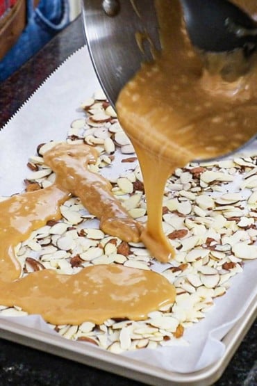 Warm toffee being poured from a metal bowl onto sliced almonds spread over parchment paper on a silver baking pan.
