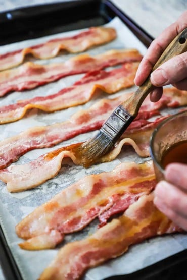 A person using a pastry brush to apply maple syrup over the tops of strips of thick-cut baking sitting on parchment paper on a baking pan.