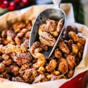 A close-up view of a person holding a metal scooper filled with spiced nuts over a Christmas tin filled with more of the nuts.