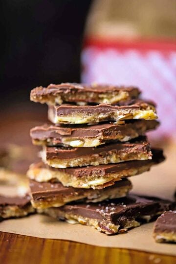 A close-up view of a stack of square chunks of chocolate toffee with almonds sitting on brown wax paper.