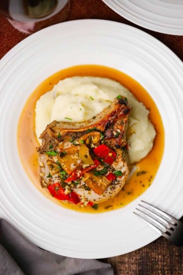 An overhead view of an Italian-style smothered pork chop resting on a bed of mashed potatoes with a pool of sauce in the bottom of the plate.