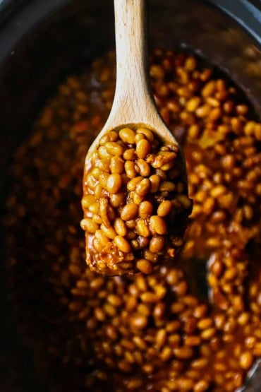 An overhead view of a large wooden spoon holding a big helping of slow cooker baked beans over the slow cooker pot filled with the same.