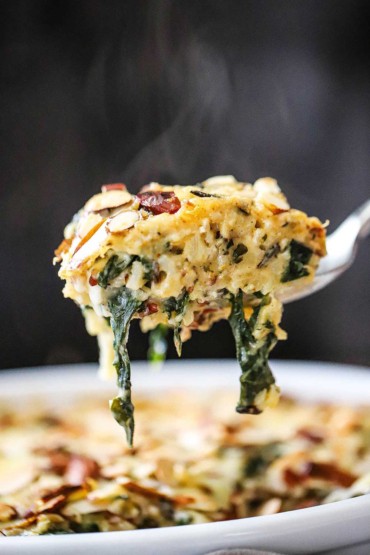 A close-up view of a large spoon lifting up a steaming helping of wild rice, spinach, and fontina casserole.