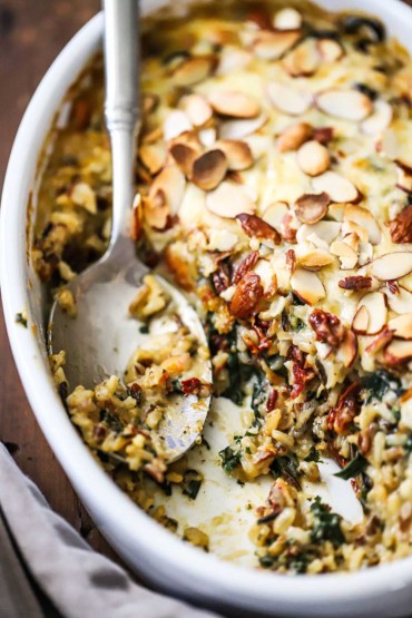 A straight-on view of an oval white baking dish partially filled with a wild rice, spinach, and fontina casserole and a serving spoon sitting in the open area of the dish.
