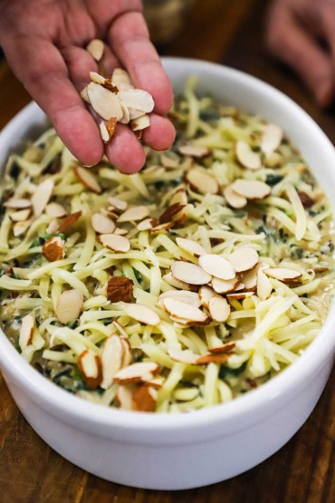 A person sprinkling almond slivers over the top of an oval baking dish that is filled with wild rice, spinach, and fontina cheese.