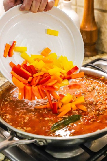 A person dumping strips of red, yellow, and orange bell peppers into a skillet filled with a simmering sauce with ground turkey.