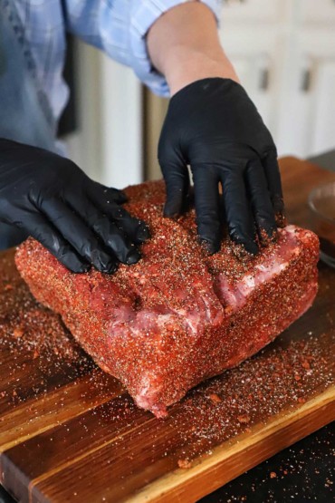 A person wearing black kitchen gloves pressing a pork rub into a large Boston butt on a wooden cutting board.