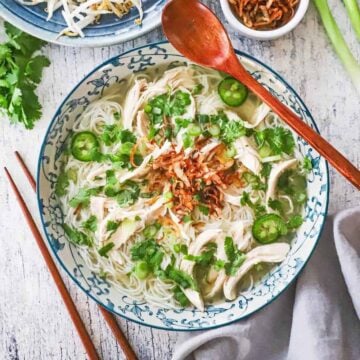 An overhead view of a colorful bowl filled with chicken pho that is garnished with sliced jalapeños, cilantro, and fried shallots.