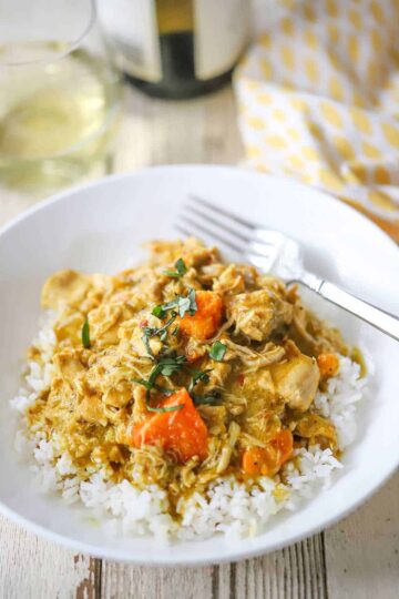 A straight-on view of a white bowl filled with a serving of slow-cooker coconut chicken curry with a glass of white wine nearby.