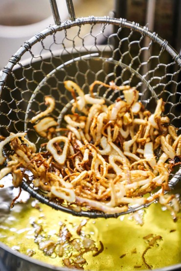 A close-up view of a metal spider holding up about a cup of thinly sliced shallot that has been fried until golden.