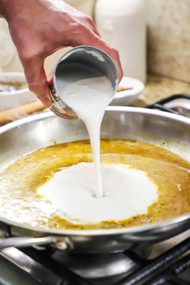 A person pouring unsweetened coconut milk from a can into a large skillet that iss filled with a thin curry sauce.