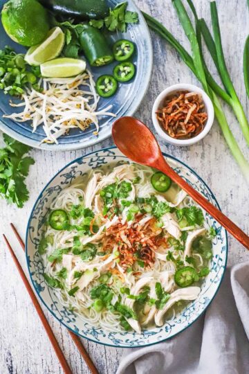 An overhead view of a bowl of chicken pho garnished with cilantro, peppers, and fried shallots, sitting next a platter of additional garnishes, including lime wedges and mung bean sprouts.