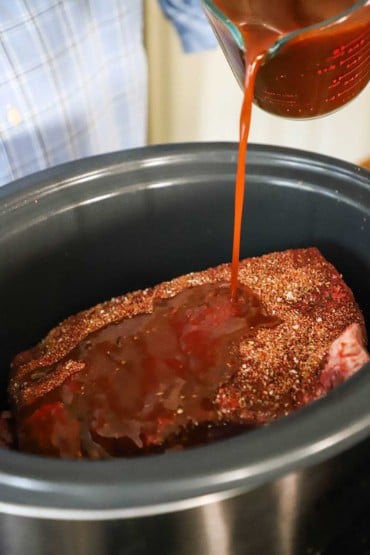 A person pouring a barbecue sauce mixture over a large seasoned brisket that is sitting in a large slow cooker.