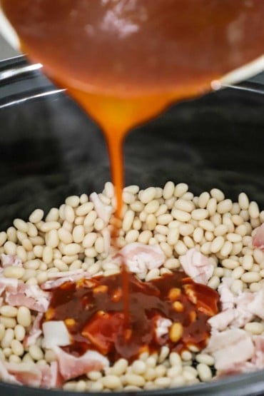 BBQ sauce being poured from a saucepan into a slow cooker filled with dried navy beans and pieces of cut-up uncooked bacon.