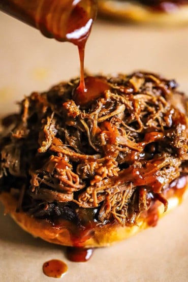 Homemade barbecue sauce being drizzled out of a glass bottle onto chopped brisket that is sitting on a toasted bun.
