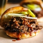 A close-up view of a slow cooker chopped brisket sandwich with barbecue sauce drizzled over the top of the meat with a couple of pickle slices and a thinly slice of onion on top, too.