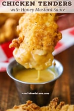 A close-up view of a crispy chicken tender that has been dipping in a small container of homemade honey mustard dipping sauce a is dripping from the bottom of the tender.