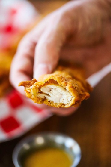 A person holding a half-eaten crispy chicken tender over a small-container of homemade honey mustard dipping sauce.