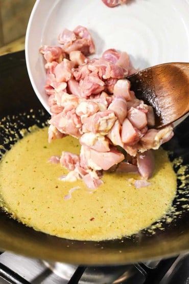 Chunks of uncooked chicken thighs being transferred from a white bowl into a large wok filled with a green curry and coconut milk sauce.