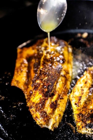 A person using a spoon to drizzle melted butter over a blackened redfish that is being cooked in a large black cast-iron skillet.