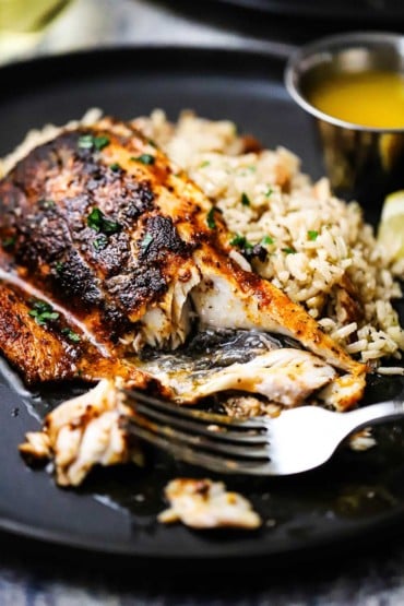 A close-up view of a half-eaten blackened redfish sitting on a black dinner plate also filled with rice pilaf and small vessel of melted butter.