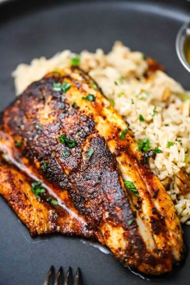 A close-up view of a blackened redfish filet resting on a bed of rice pilaf on a black dinner plate.